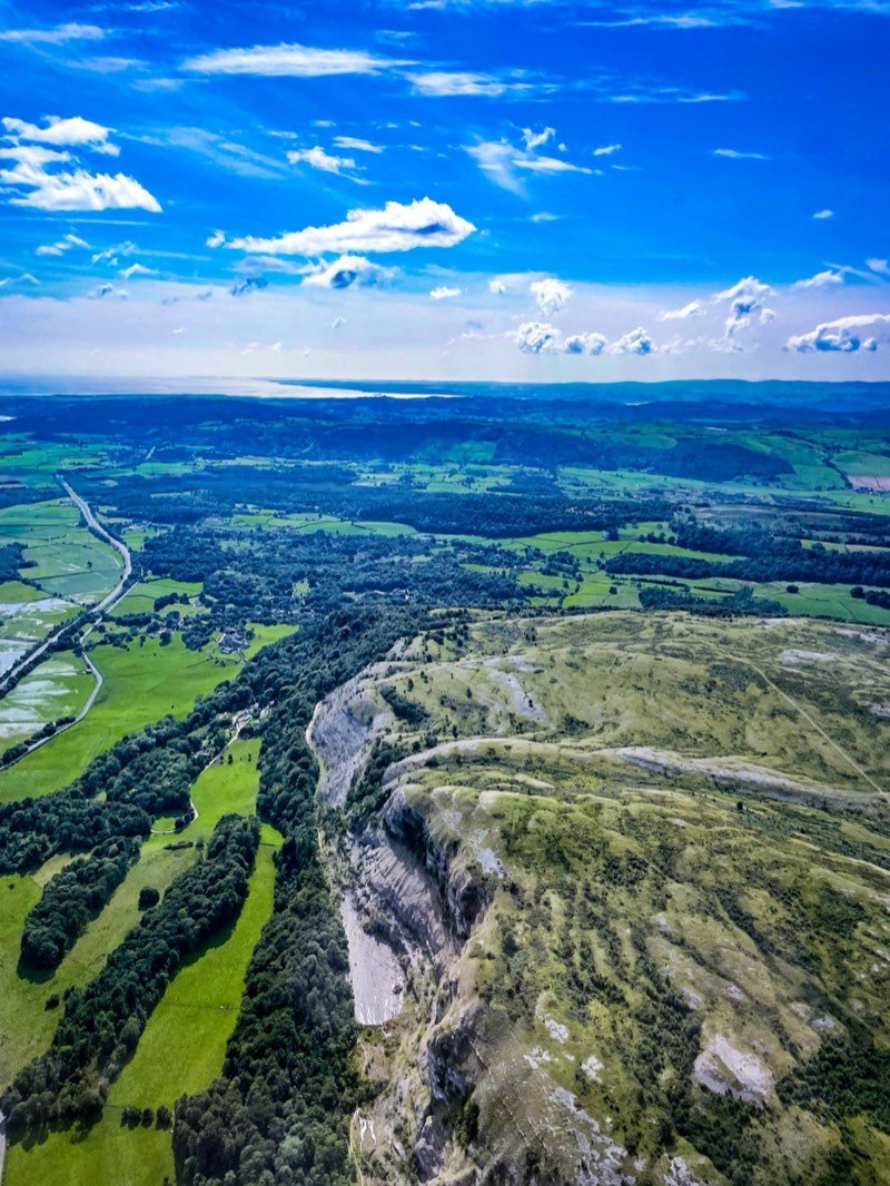 A view from the air along a pale limestone escarpment, with green fields and woodland below and the coast on the horizon.