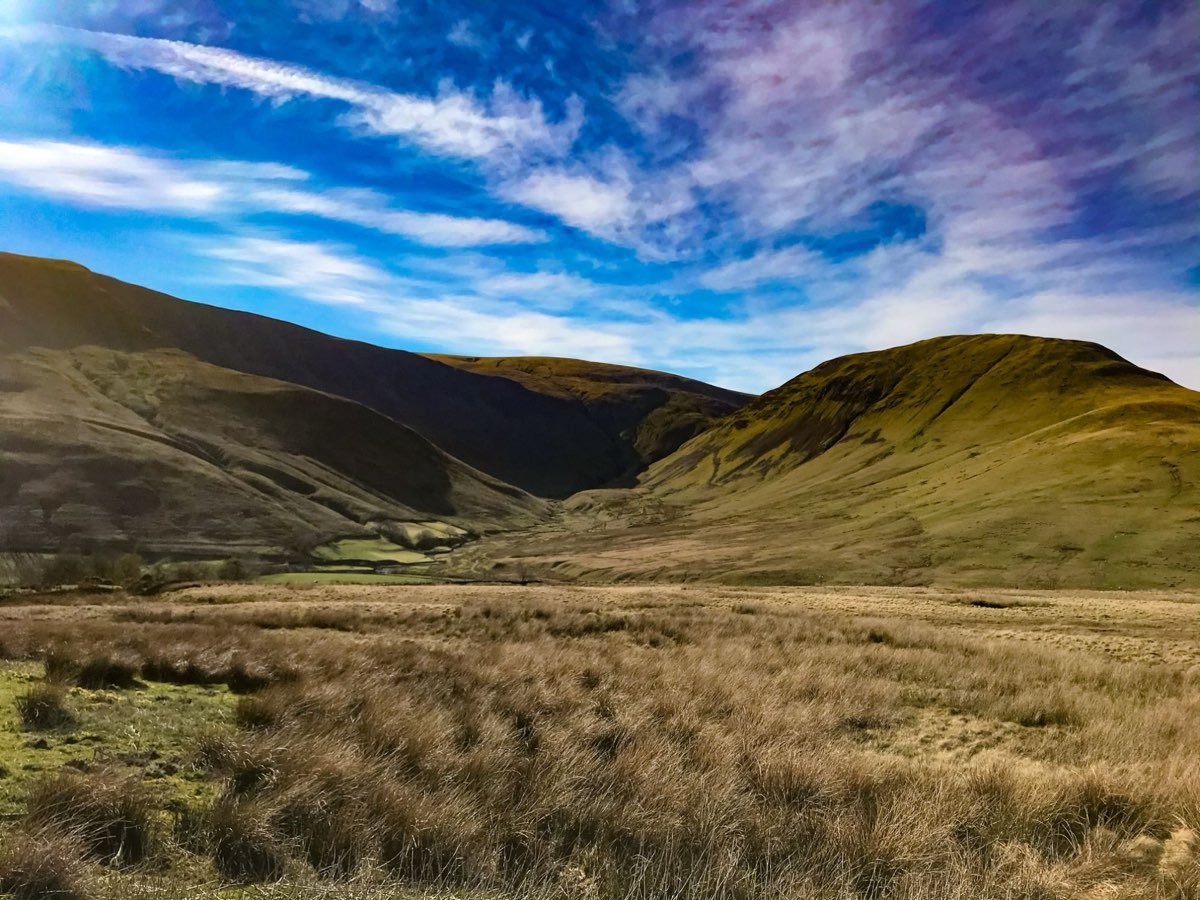 The mouth of a wide grassy valley between two smooth fells under a blue sky streaked with cloud, with rough moorland grass in the foreground.