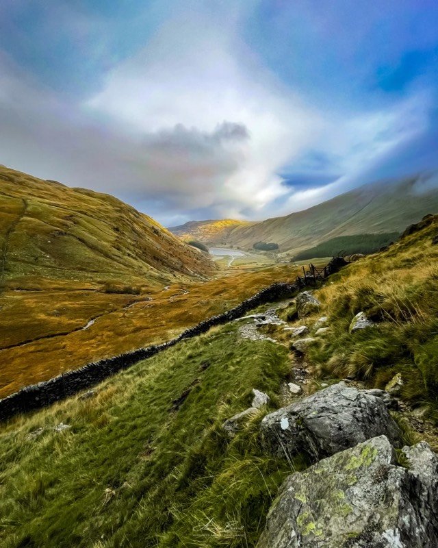 A rocky path and dry-stone wall running down a grassy valley towards a reservoir, under a bright, cloudy sky.