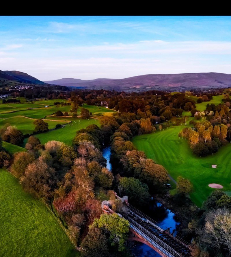 An iron arch bridge crossing a river beside a golf course among autumn trees, seen from the air, with hills in the distance.
