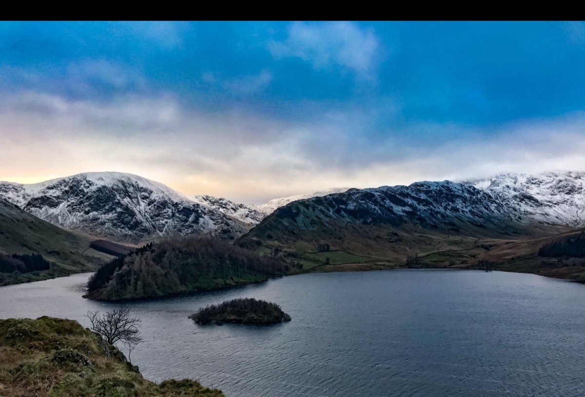 Snow-dusted fells above a dark lake with a wooded promontory and a small island under a blue and gold sky.