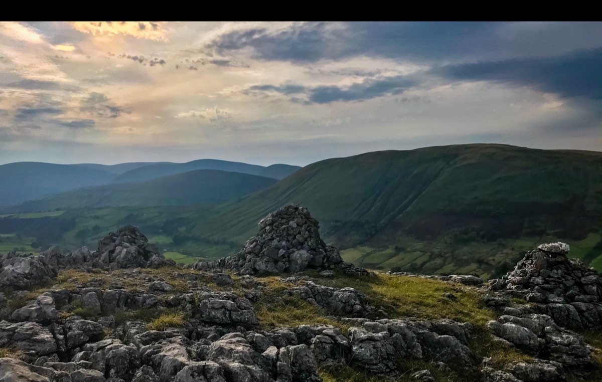 Stone cairns on rough limestone in the foreground, with a long green fell and a hazy valley behind under a dramatic sky.