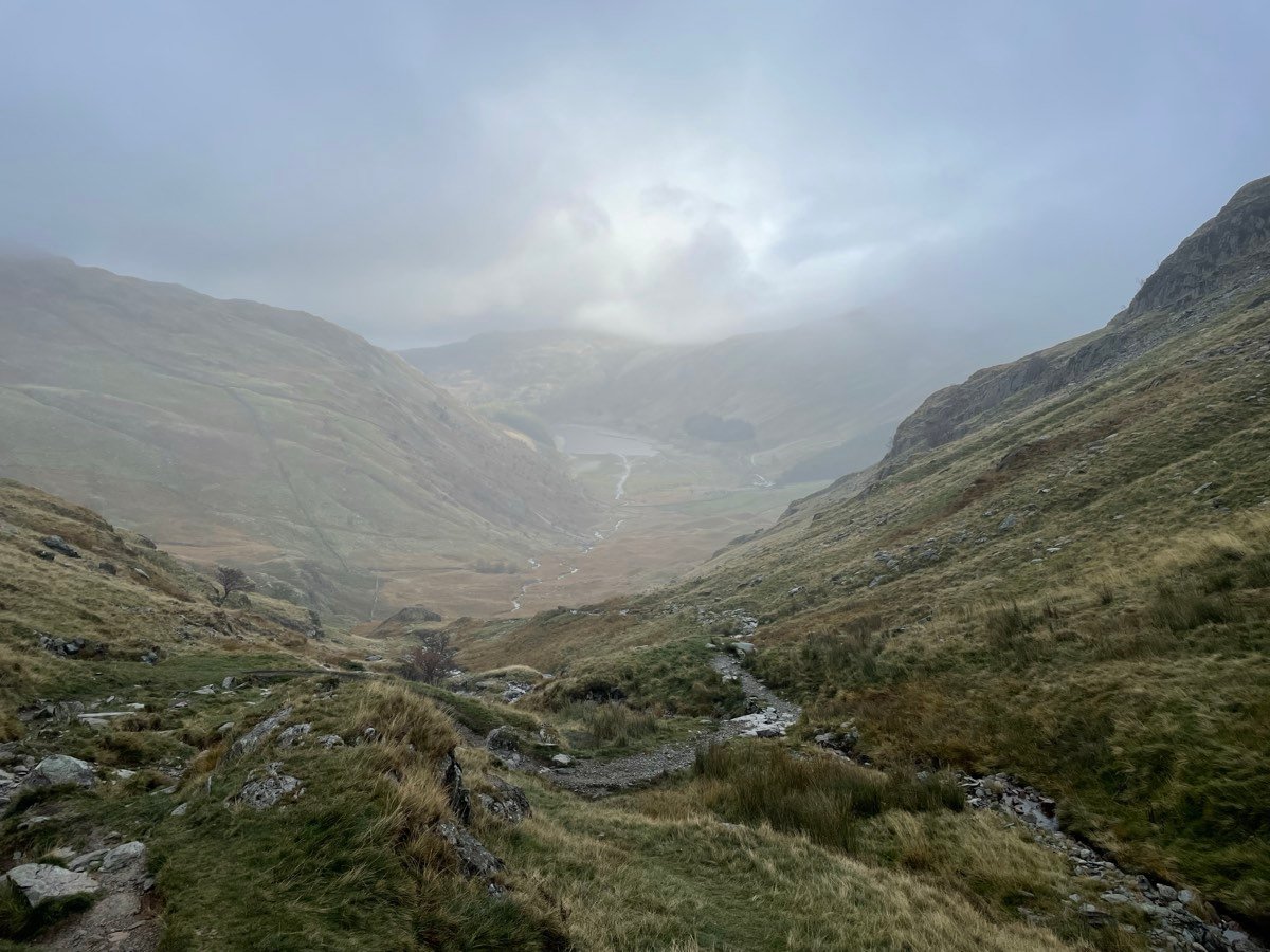 A rocky path dropping into a misty valley between steep green hillsides.