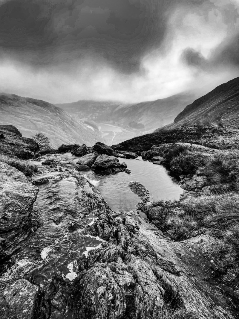 A black and white view over a rocky pool towards a deep valley with a reservoir far below and low cloud.