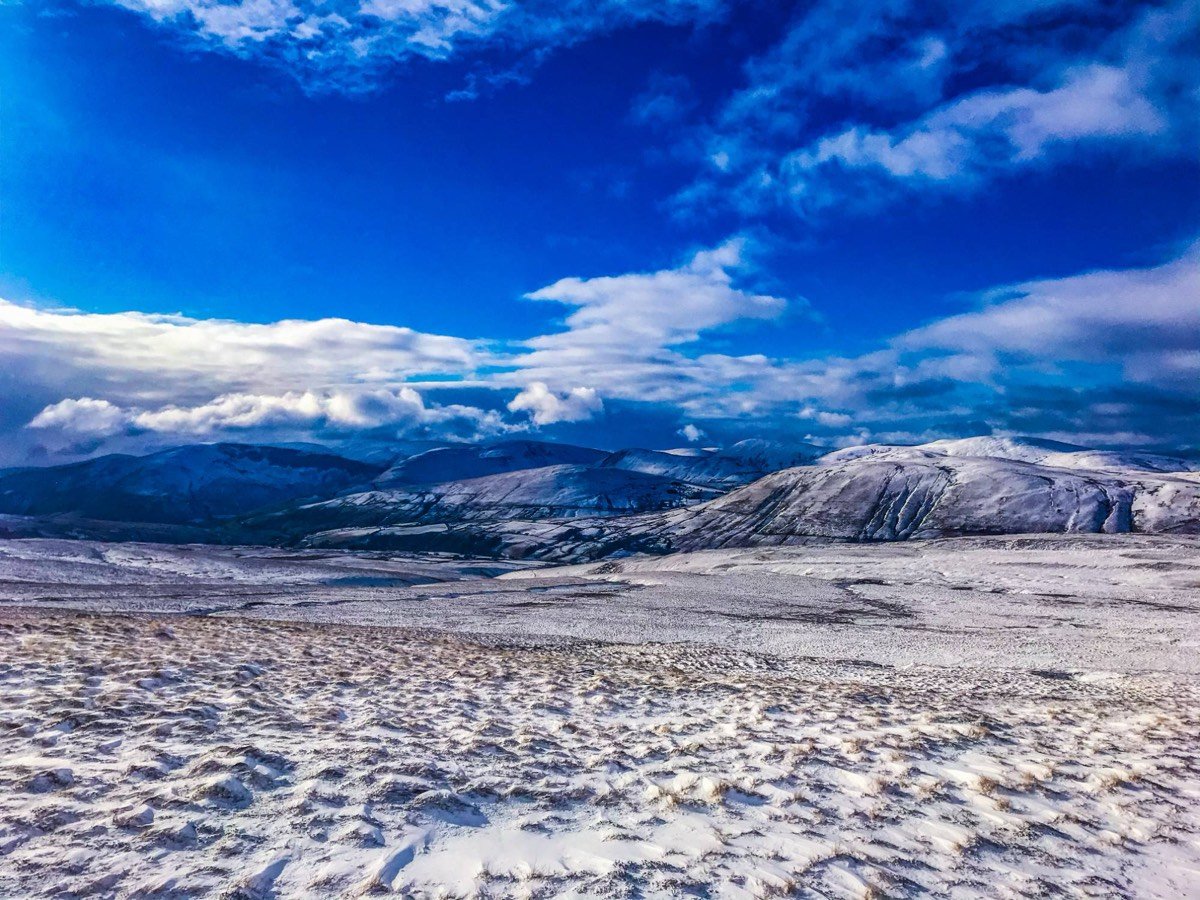 A wide snow-dusted upland with white-capped fells in the distance under a deep blue sky with patchy cloud.