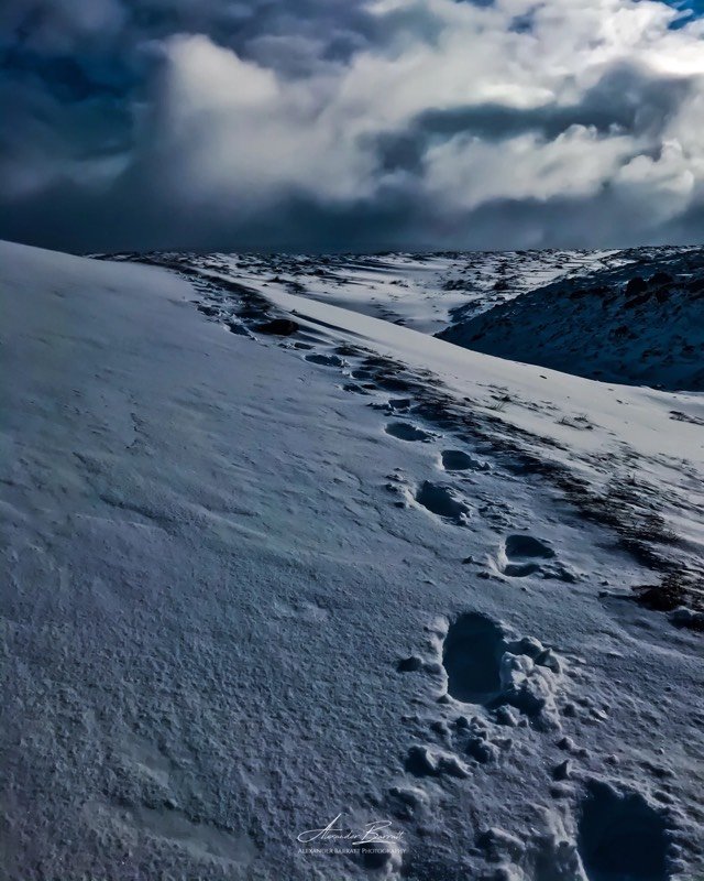 Footprints leading up a snow-covered slope towards a dark, stormy sky.