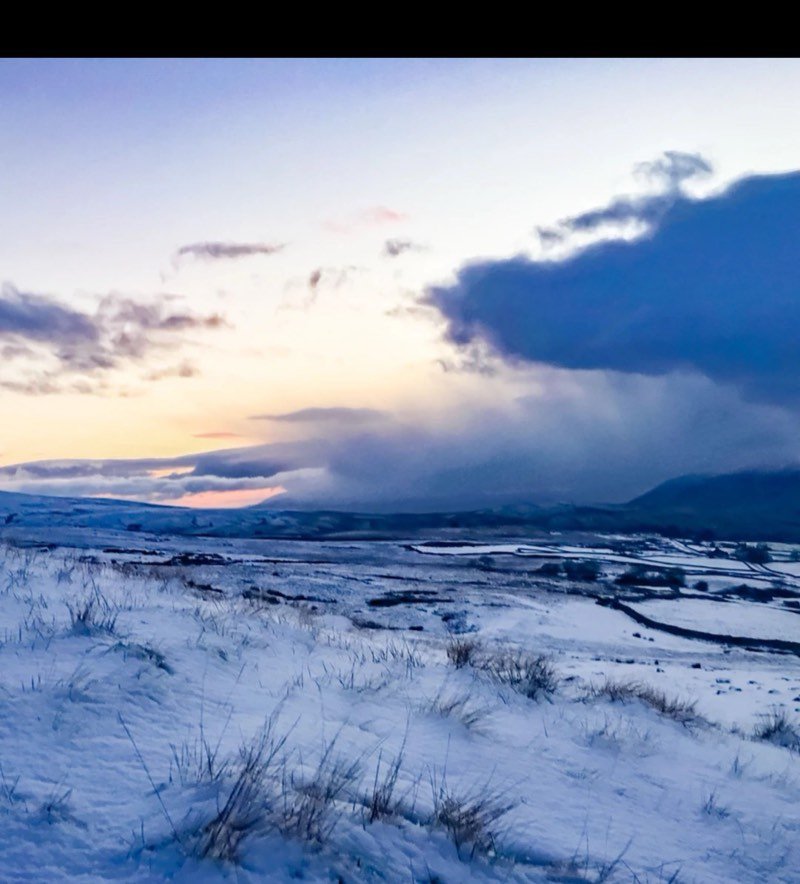 Fresh snow on open moor with a dark storm cloud rolling in under a pale evening sky.