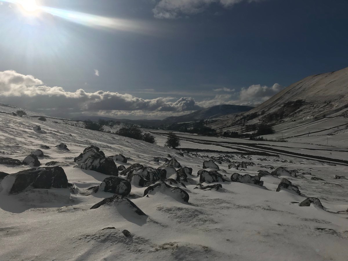 Dark boulders and drifted snow across a wide dale, with a low sun and hills behind.