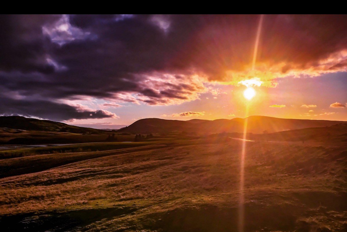 The sun bursting through purple storm cloud above dark fells and a field of golden grass.