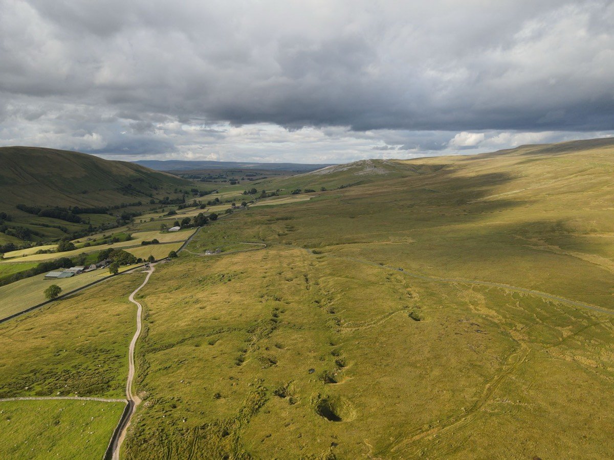 A drone view down a green valley with a farm and a winding track, and a road crossing open moorland under heavy grey cloud.