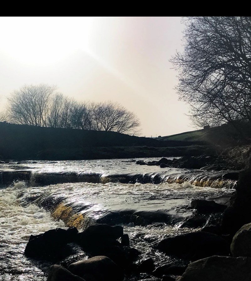 Backlit water tumbling over a stone weir between dark rocks, with bare trees against a pale sky.